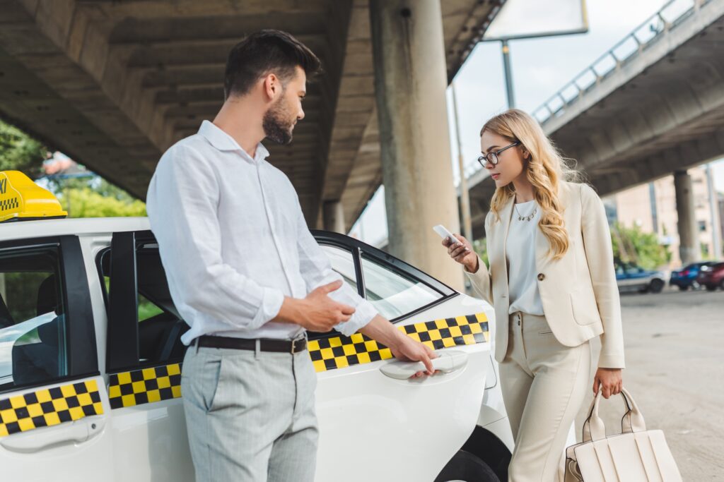 young man opening door of taxi to beautiful woman in eyeglasses using smartphone